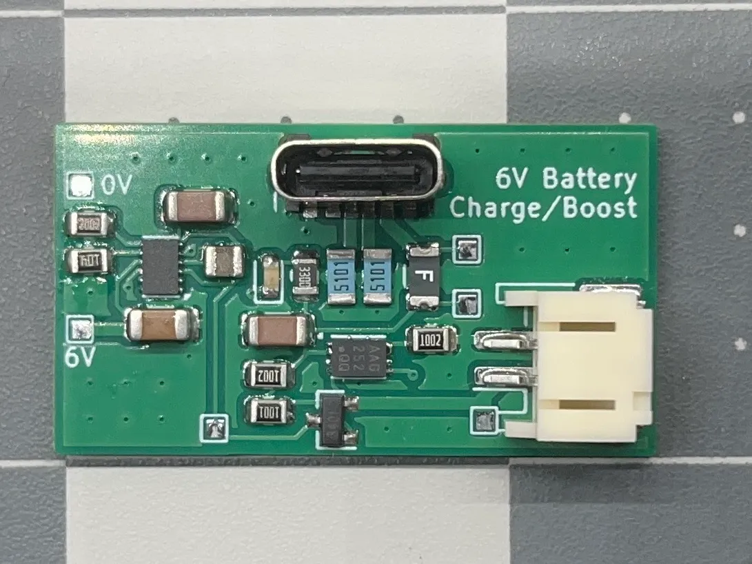 A top-down picture of a green PCB containing two large chips and several small passives. On the left, there are two small pads labeled 0V and 6V. In the middle near the top is a USB C port facing the camera. In the top right there is text printed on the PCB: "6V Battery Charge/Boost." In the bottom right there is a socket for a JST-SH connector, pointing to the right.